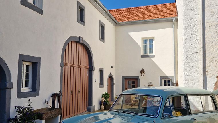 Une voiture classique, verte menthe, est garée devant un bâtiment charmant et lumineux. Le ciel est clair et bleu, ce qui crée une atmosphère accueillante.