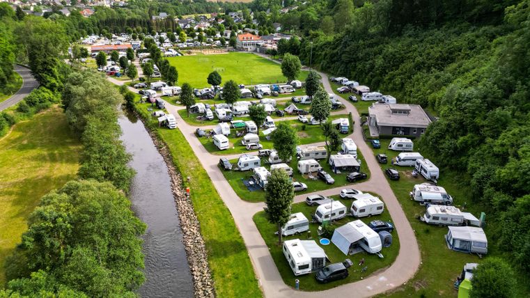 Ein schöner Campingplatz neben einem Fluss, umgeben von grüner Natur. Viele Wohnmobile und Zelte sind auf dem Platz verteilt.