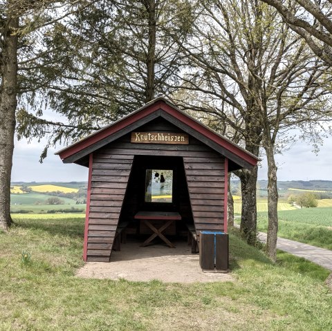 Een kleine houten hut met het bordje 'Knutschheisjen' staat tussen bomen in een groen landschap van velden en heuvels., © A. Girards