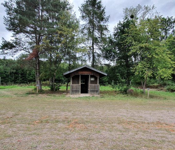 Kleine houten hut in het bos, omringd door bomen en een grasveld. De lucht is bewolkt., © TI Bitburger Land - Steffi Wagner