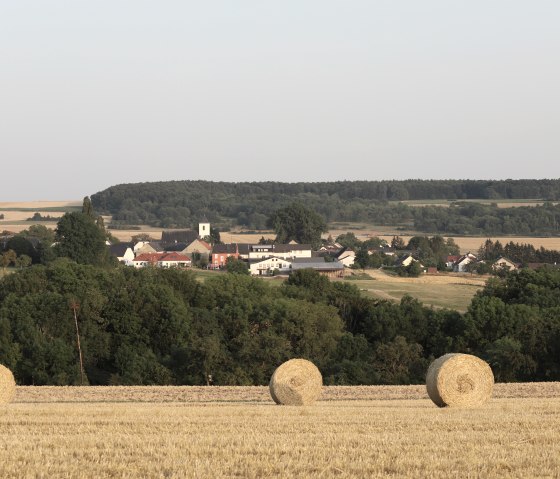 View of a village with a church, surrounded by fields and woods. In the foreground, several bales of straw lie on a harvested field., © Irmgard Rüdiger