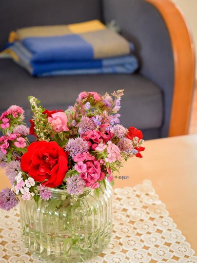 Ein schöner Blumenstrauß mit roten und rosa Blumen in einer klaren Glasvase. Im Hintergrund sind ein Sofa und ein gefalteter Überwurf zu sehen.
