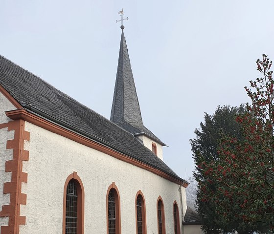 Eine Kirche in Idesheim mit einem Kreuz an der Fassade und einem Baum mit roten Beeren im Vordergrund., © TI Bitburger Land