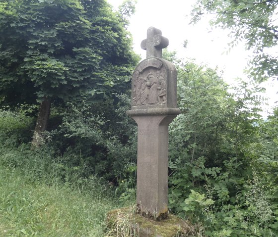 Ein steinernes Kreuzwegdenkmal steht in einer grünen, bewaldeten Umgebung. Es zeigt eine Reliefdarstellung und ist von Bäumen umgeben., © TI Bitburger Land