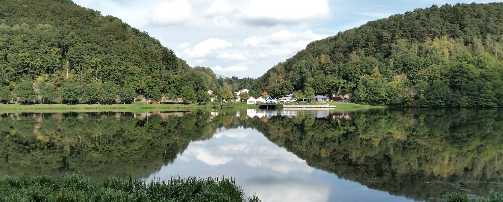 Panorama van het Bitburg stuwmeer met beboste heuvels, gebouwen aan de oever en een heldere weerspiegeling in het water onder een blauwe hemel., &copy; TI Bitburger Land