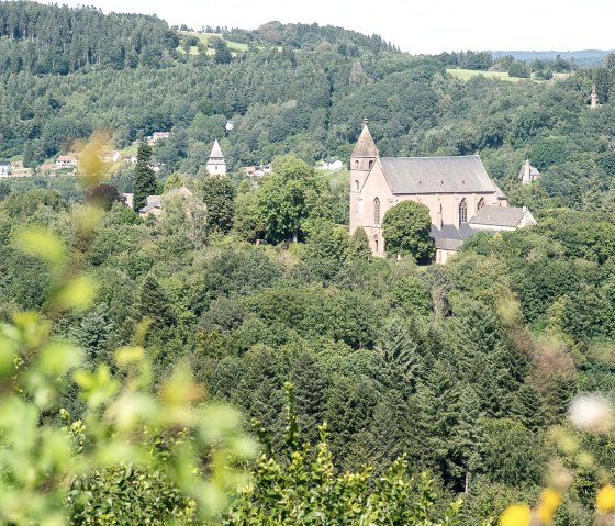 Blick auf eine Kirche, umgeben von dichten Wäldern und Hügeln, unter blauem Himmel. Im Vordergrund sind unscharfe Pflanzen zu sehen., © Tourist-Info Bitburger Land M.Mayer