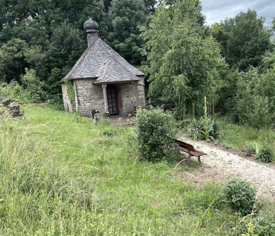 Une petite chapelle en pierre avec un toit en ardoise se dresse au milieu d'une végétation luxuriante. Un chemin de gravier mène à un banc en bois devant la chapelle., © Daniel Köhler