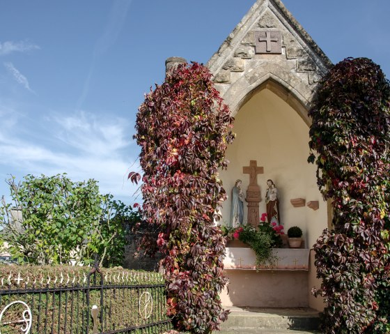 Kleine Kapelle mit Statuen, umgeben von herbstlichem Efeu. Ein schmiedeeisernes Tor und blauer Himmel im Hintergrund., © TI Bitburger Land