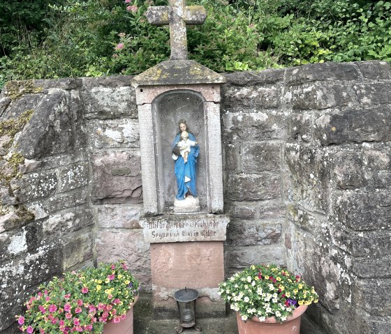 Ein steinernes Wegekreuz mit einer Marienfigur in einer Nische, flankiert von zwei Blumentöpfen, vor einer Steinmauer in Oberweis., © Daniel Köhler