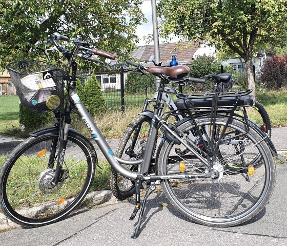 Zwei Fahrräder stehen an einer Straße in Ingendorf. Ein Wegweiser zeigt Richtung Bettingen. Im Hintergrund sind Bäume und Häuser zu sehen., © TI Bitburger Land, Steffi Wagner