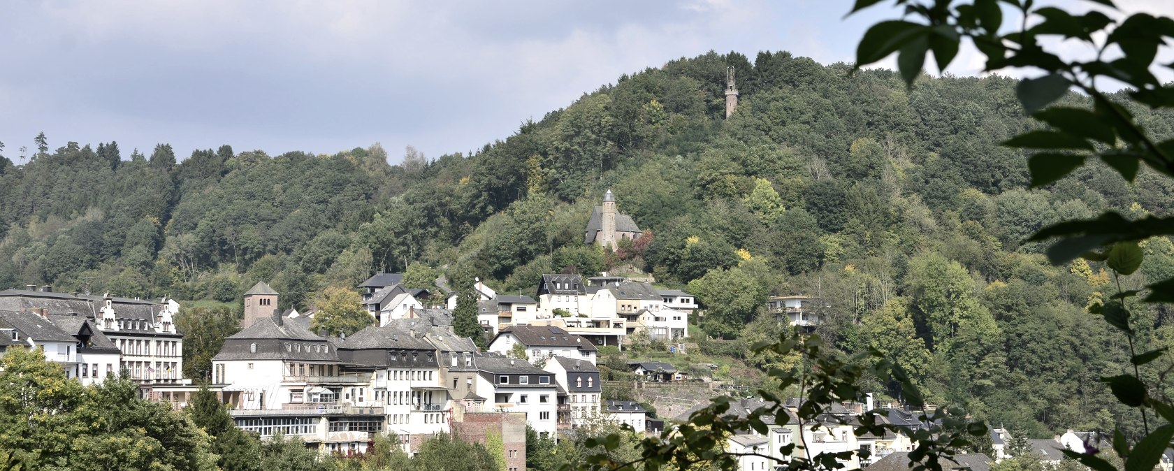 Panoramablick auf Kyllburg mit Häusern und einer Kirche auf einem bewaldeten Hügel unter blauem Himmel., © TI Bitburger Land