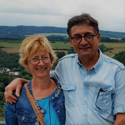 Un couple souriant pose devant un paysage pittoresque. En arrière-plan, on peut voir des collines verdoyantes et un ciel nuageux.