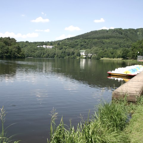 Le lac de barrage de Bitburg aux eaux calmes, entouré de collines verdoyantes. Des pédalos sont amarrés au ponton, des gens se détendent sur la rive. Une belle journée d'été., © TI Bitburger Land