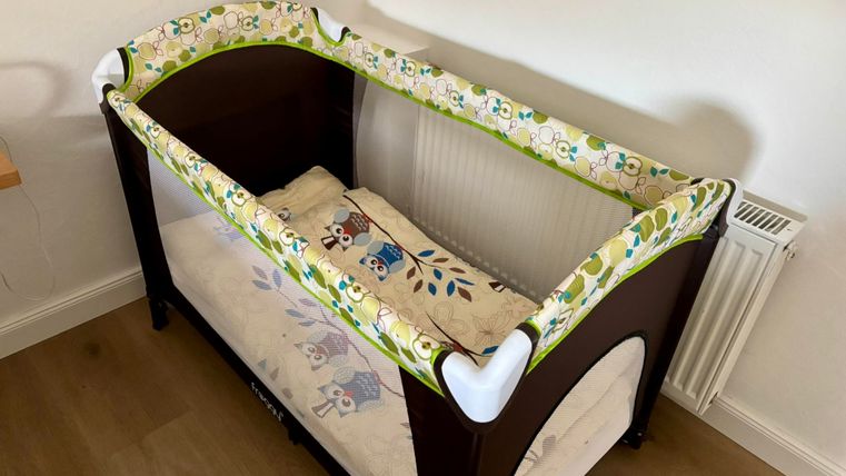 Un lit d’enfant avec un matelas coloré et des bords doux se trouve dans une pièce lumineuse. Le sol est en bois, et le lit est bien éclairé.