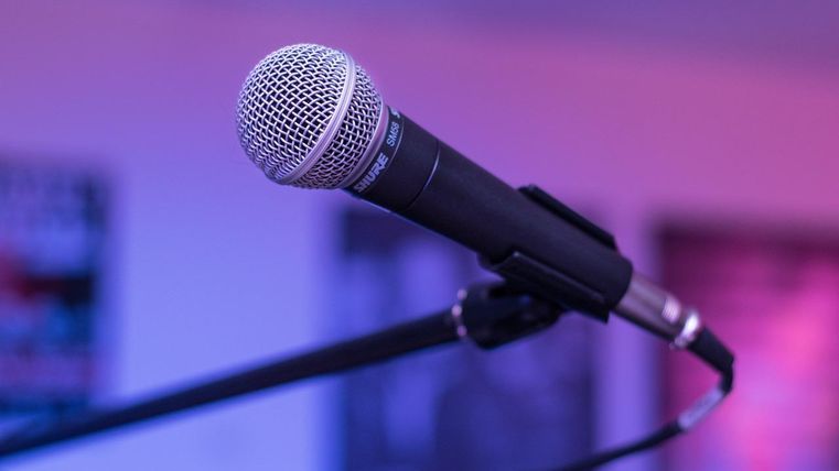 A microphone with a black body and a silver grill. In the background, blurred colors and shapes can be seen.