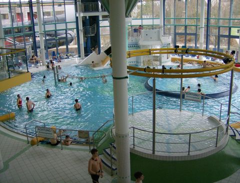 A large swimming pool with various water areas and many visitors. In the background, slides and large windows can be seen.