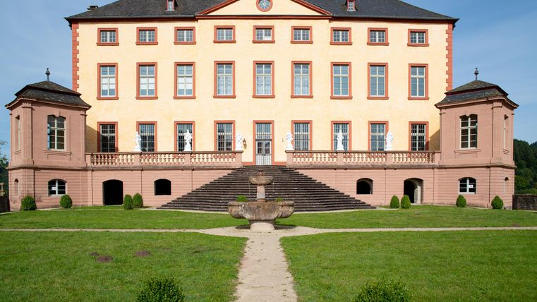 Ein elegantes Schloss mit einer hellen Fassade und großen Fenstern. Der gepflegte Garten mit einem Weg und einem Brunnen lädt zum Verweilen ein.
