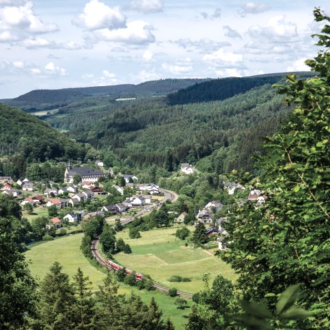 View of St. Thomas with green hills, a village in the valley and a railroad line. The sky is slightly cloudy., © TI Bitburger Land