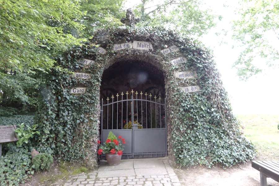 Une grotte de Lourdes recouverte de lierre avec une porte. Des fleurs sont visibles sur le portail. Des panneaux avec des inscriptions décorent l'entrée., © Eifelverein Ortsgruppe Speicher