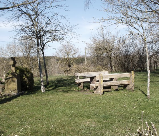 Rastplatz mit Holzbänken und steinernem Kreuz auf einer Wiese, umgeben von kahlen Bäumen unter blauem Himmel., © M. Bach
