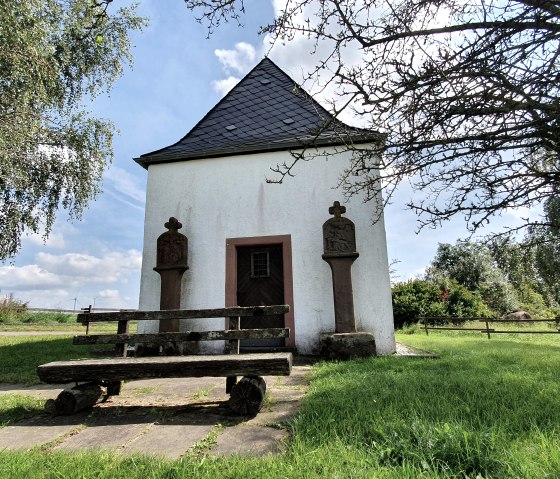 Weiße Kapelle mit Schieferdach, flankiert von zwei Steinsäulen. Eine Holzbank steht davor, umgeben von grüner Wiese und Bäumen., © Tourist-Information Bitburger Land