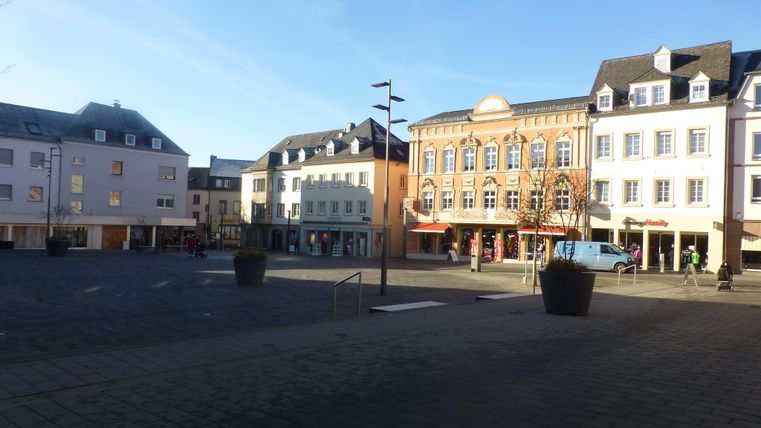 Ein ruhiger Platz mit mehreren Gebäuden im Hintergrund. Die Straße ist pflastersteingedeckt und vor den Häusern stehen große Blumentöpfe.
