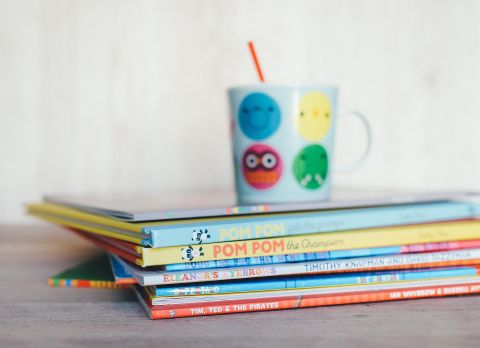 A stack of colorful children's books lies on a table. Next to it stands a cup with cheerful faces and a straw.