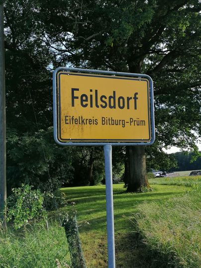 Un panneau de localité avec l'inscription "Feilsdorf" dans le district de Bitburg-Prüm. Entouré d'arbres et d'herbe.