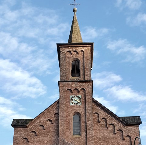 Backsteinkirche St. Laurentius mit Turm und Uhr, umgeben von Bäumen und einem Kreuz im Vordergrund, unter blauem Himmel., © TI Bitburger Land