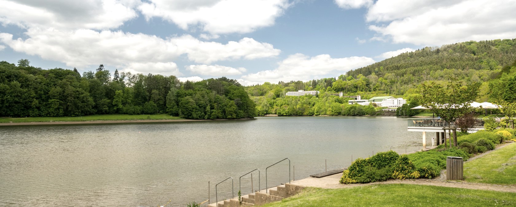 Bitburg reservoir with green shore landscape, trees and buildings in the background under a cloudy sky., © Eifel Tourismus GmbH, Dominik Ketz