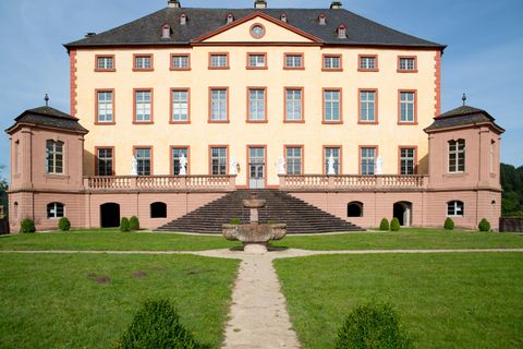 A magnificent castle with a bright yellow coat of paint and large windows. In front of the castle, there is a well-maintained garden with a path and a small fountain.