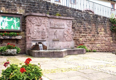 Ein steinerner Dorfbrunnen mit Reliefs und fließendem Wasser, umgeben von roten Blumen und einer Infotafel an einer Steinmauer., © TI Bitburger Land - M. Mayer