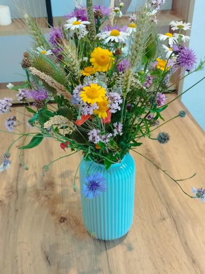 Ein bunter Blumenstrauß mit verschiedenen Wildblumen in einer blauen Vase. Der Strauß steht auf einem Holztisch.