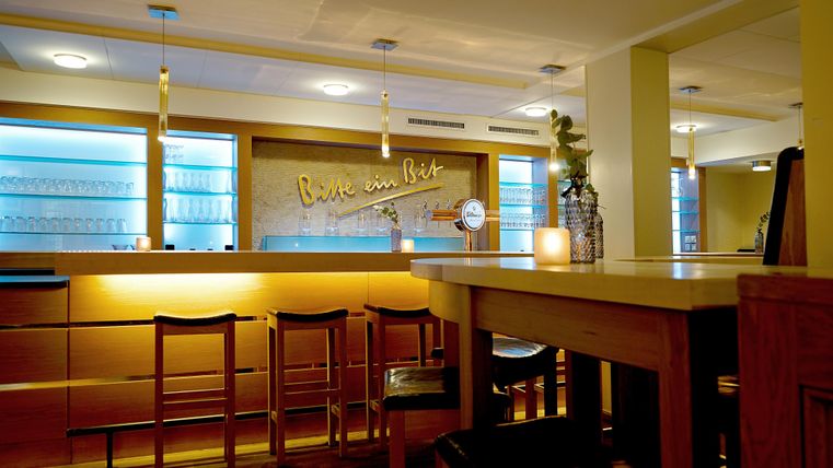 Ein gemütliches Restaurant mit einer eleganten Bar und warmem Licht. Die Wände sind in hellen Farben gestaltet, und es gibt einige Pflanzen für einladende Atmosphäre.