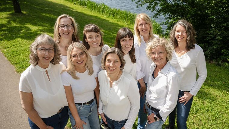 Een groep vrouwen in formele kleding staat voor een fontein in een park. Op de achtergrond zijn meerdere vlaggen en gebouwde structuren zichtbaar.