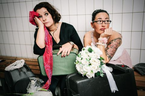 Twee vrouwen zitten op koffers en kijken mijmerend. Een van hen houdt een boeket vast, terwijl de ander een sjaal draagt.