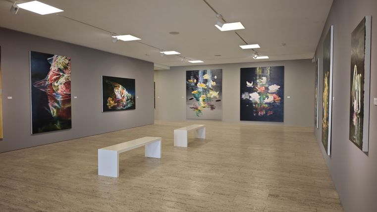 Ein moderner Ausstellungsraum mit mehreren bunten Kunstwerken an den Wänden. In der Mitte steht eine weiße Bank auf dem Boden.