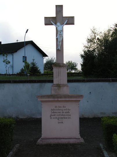 Stenen kruis met Jezusfiguur op een begraafplaats, omringd door bomen en een huis op de achtergrond.