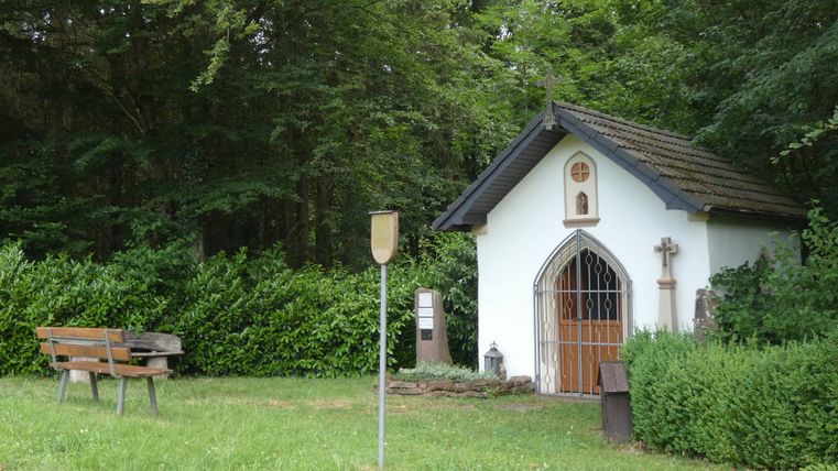 Kleine Kapelle im Grünen mit Bank und Schild.