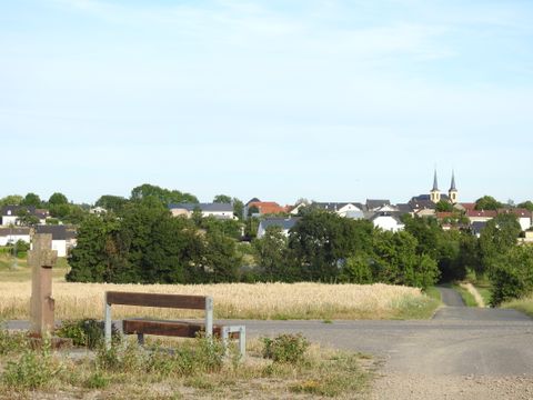 Landschaft mit Bank, Feld und Dorf im Hintergrund.