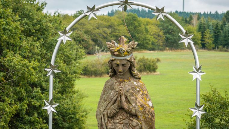 Une statue d'une sainte entourée d'un arc métallique avec des étoiles. En arrière-plan, un paysage verdoyant s'étend et des éoliennes sont visibles.