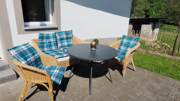 Ein gemütlicher Sitzbereich im Freien mit einem runden Tisch und drei Korbsesseln. Die Sessel sind mit blauen und grünen Kissen bezogen.