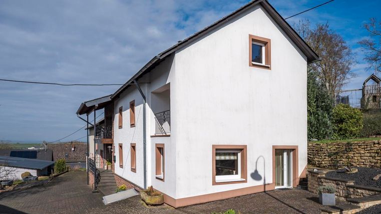Ein modernes Haus mit heller Fassade und roten Akzenten. Es steht an einer ruhigen Straße und ist von einer grünen Umgebung umgeben.