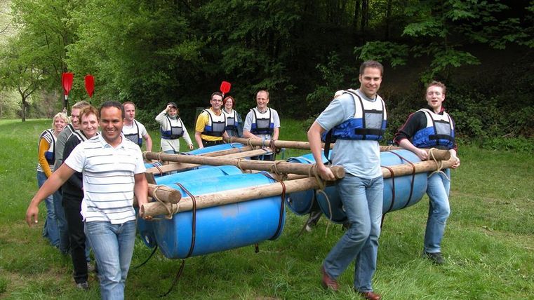 Eine Gruppe von Menschen trägt ein selbstgebautes Floß über eine Wiese. Im Hintergrund sind Bäume und ein Brückenbogen zu sehen.