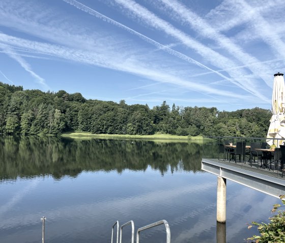 Bitburg reservoir with terrace and parasol, surrounded by forest. The sky is blue with vapor trails., © TI Bitburger Land