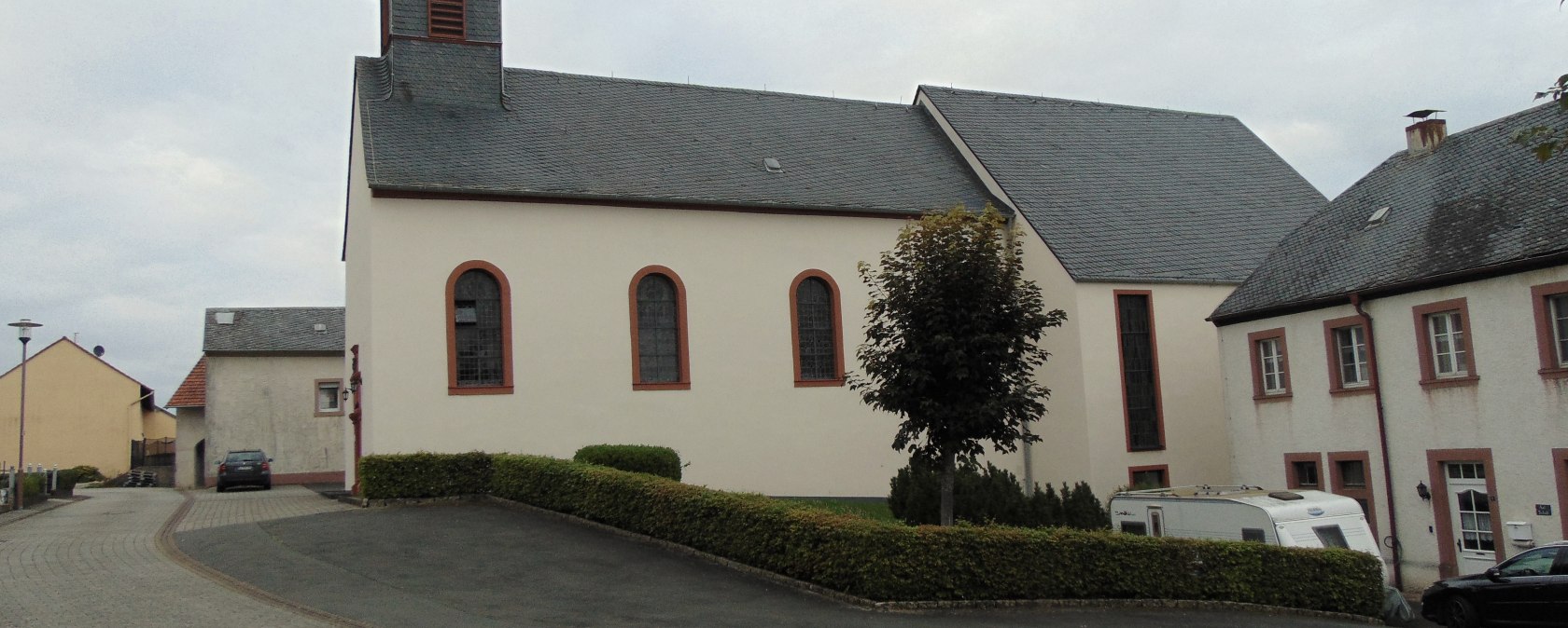 De kerk St. Urban in Gindorf met een spitse toren, omgeven door huizen en een geplaveid pad. Een boom en een caravan zijn zichtbaar., © Conny Meier