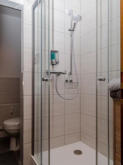 Eine moderne Dusche mit Glasschwingtüren und weißen Fliesen. Im Hintergrund ist eine Toilette sichtbar.