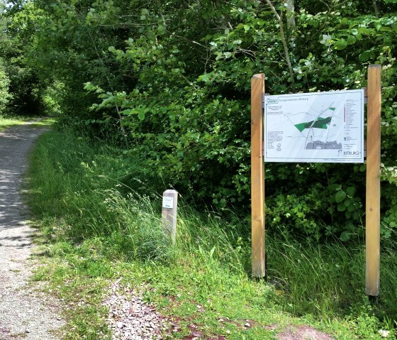 A shady forest path leads through lush greenery. On the right is a large information sign with a map. The path is lined with trees., © Tourist-Information Bitburger Land