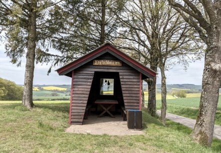 Eine kleine Holzhütte mit dem Schild 'Knutschheisjen' steht zwischen Bäumen in einer grünen Landschaft mit Feldern und Hügeln., © A. Girards