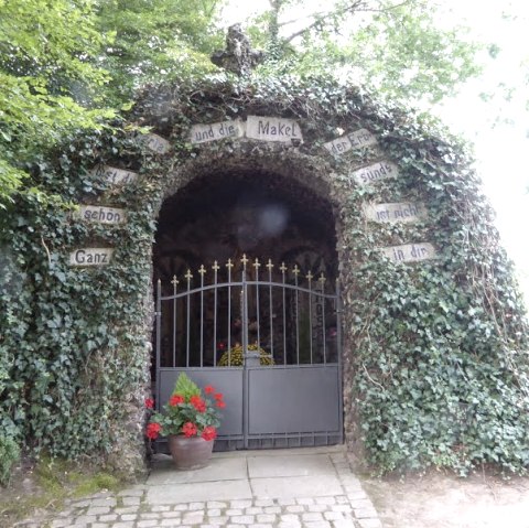 Een met klimop begroeide Lourdesgrot met een poort. Op de poort zijn bloemen te zien. Borden met letters sieren de ingang., © Eifelverein Ortsgruppe Speicher